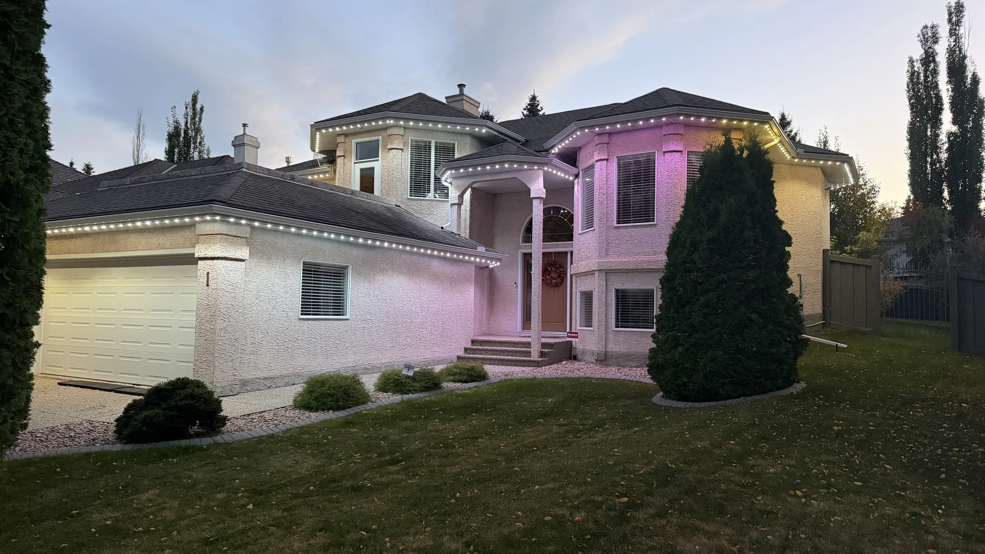 Side-angle view of a home with white lights tracing its garage and upper eaves and purple accents near the front entry